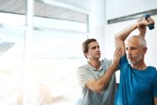 Trainer korrigiert die Haltung eines älteren, grauhaarigen Patienten in blauem T-Shirt , der Übungen mit einer Kleinhantel macht Trainer korrigiert die Haltung eines älteren, grauhaarigen Patienten in blauem T-Shirt , der Übungen mit einer Kleinhantel macht